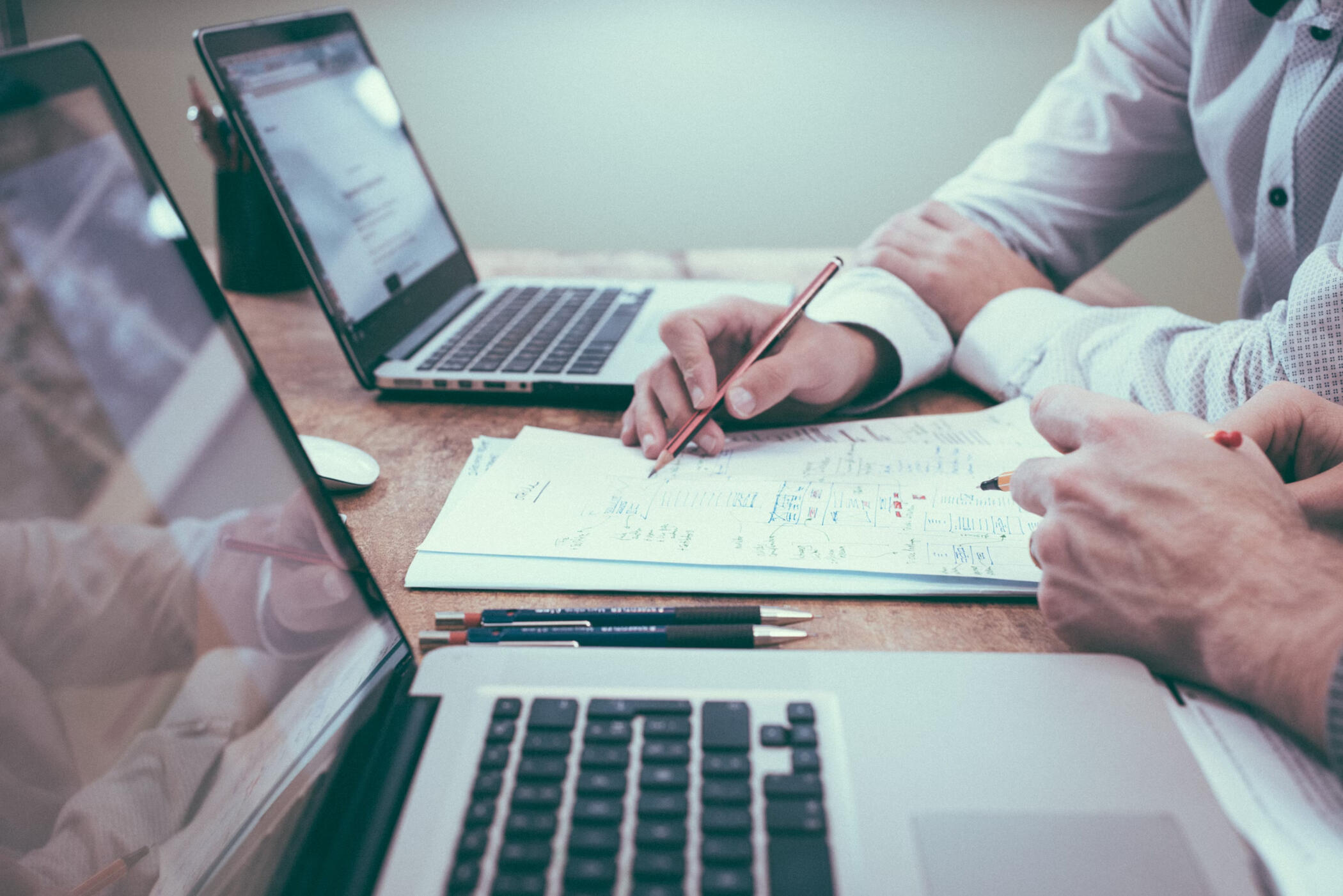 Close-up of two professionals working together at a desk, reviewing documents and notes with laptops open. One person is writing on paper, while the other points at information, illustrating teamwork and strategic business planning.