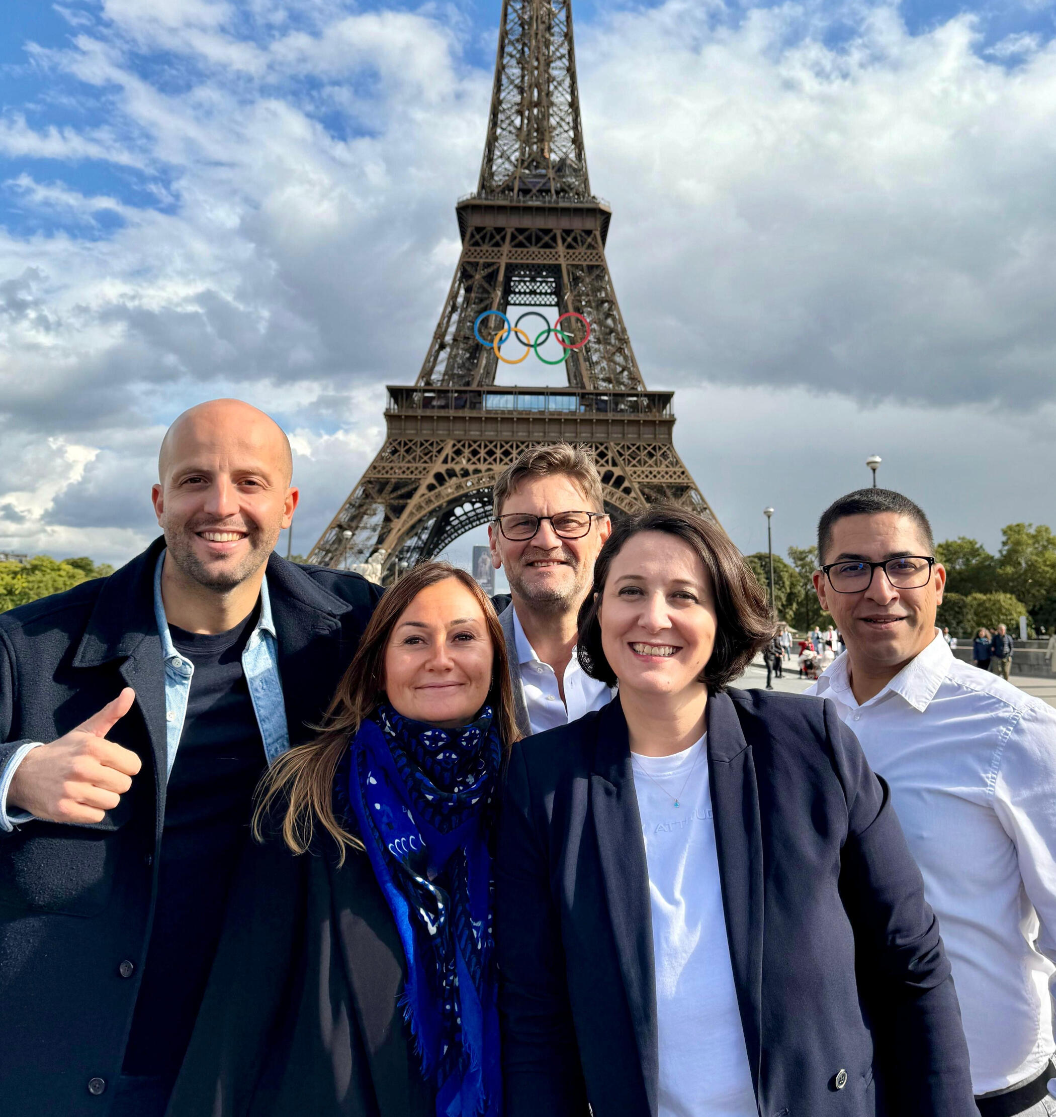 A group of five smiling professionals from the Hahn Zekker team standing in front of the Eiffel Tower, with Olympic rings displayed on the tower and a partly cloudy sky in the background.