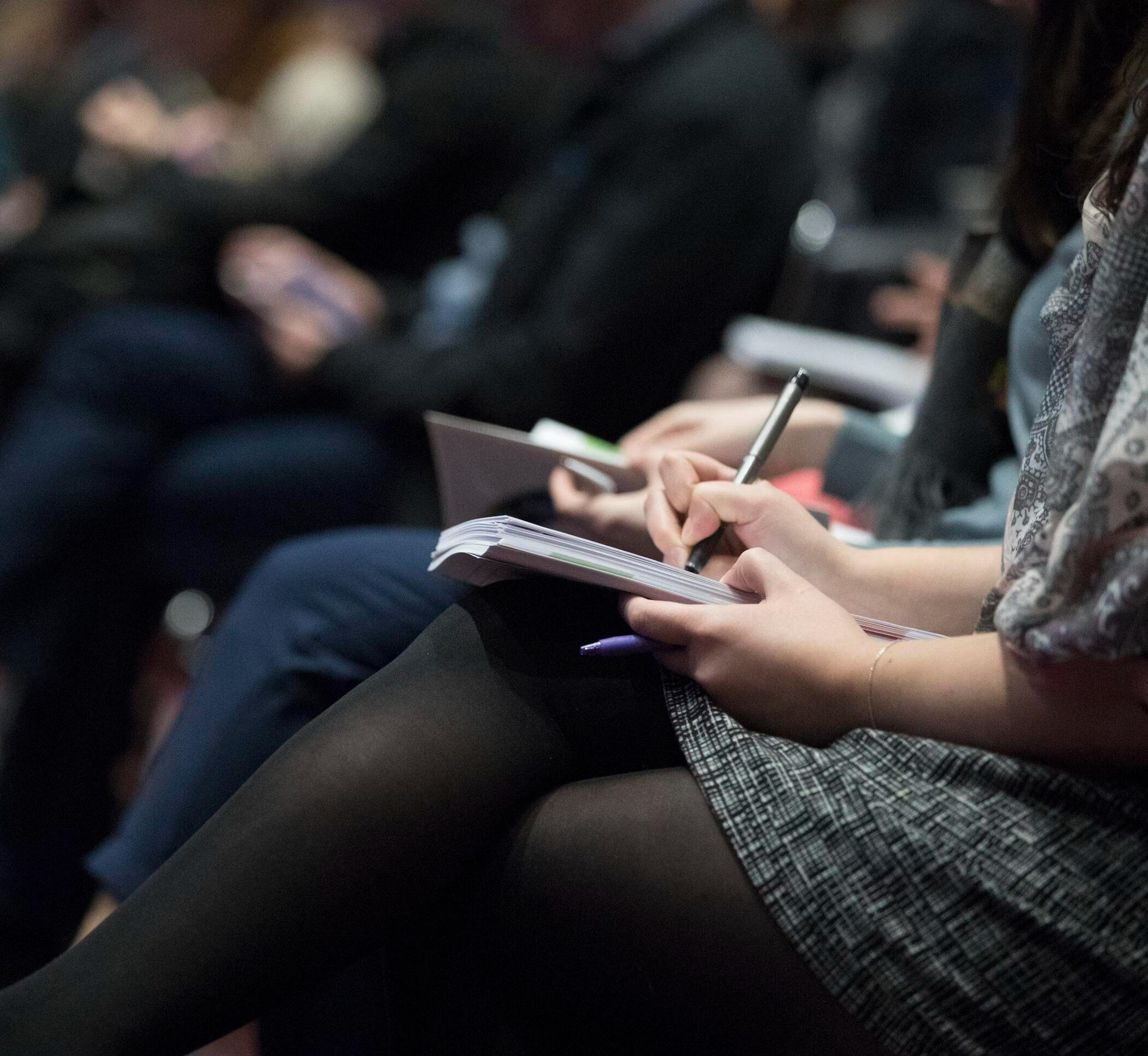 Close-up of professionals seated at a seminar or conference, focused on a woman writing notes in a notebook. The scene captures active participation and engagement during a business or leadership event.