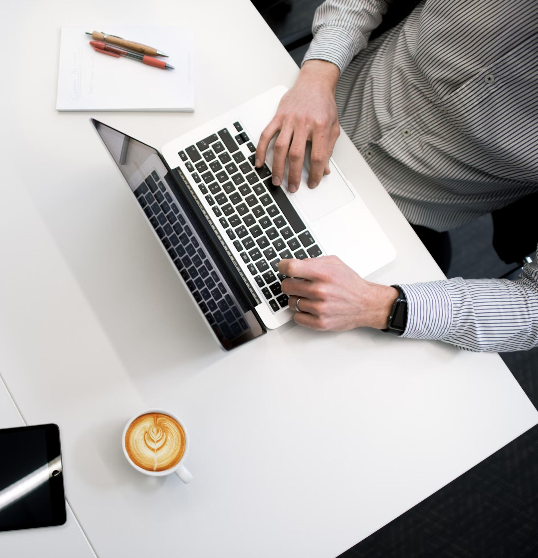 Top-down view of a business professional working on a laptop at a clean white desk, with a notepad, coffee, conference phone, remote control, and tablet in view.
