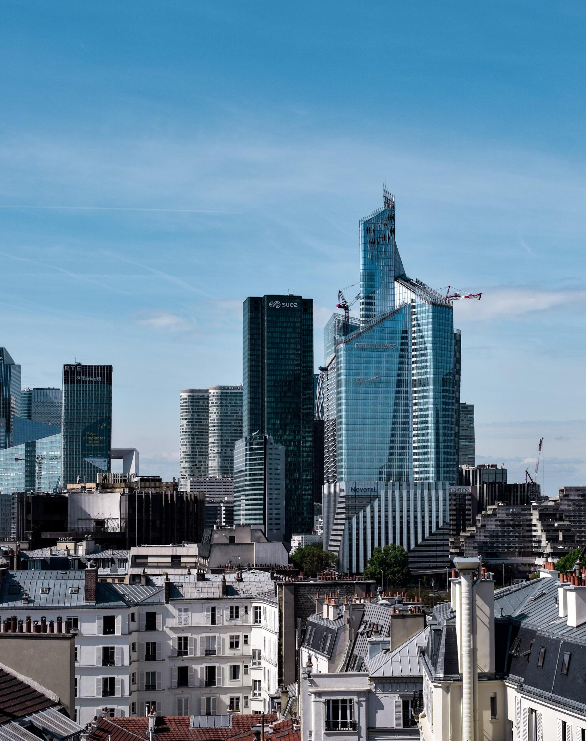 A panoramic view of Paris blending classic Parisian rooftops in the foreground with the modern glass skyscrapers of La Défense business district in the background under a clear blue sky.