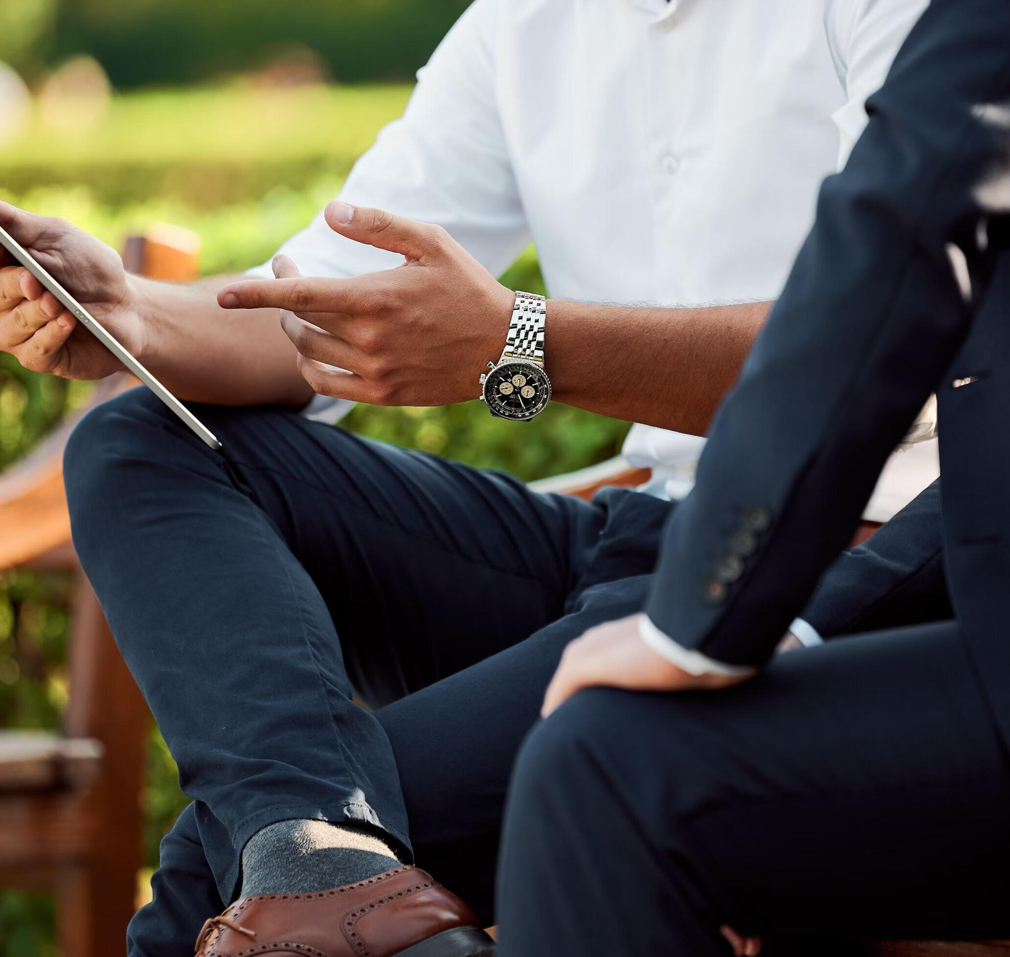 Two business professionals sitting on a bench outdoors, discussing work while looking at a tablet. One is wearing a white shirt and a silver watch, the other is in a dark suit. The setting is relaxed and professional, with greenery in the background.